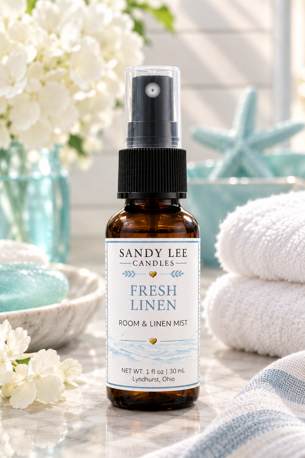 Sandy Lee Candles Fresh Linen Luxury Room & Linen Mist in an amber glass bottle styled with crisp white linens, white hydrangeas, soft turquoise accents, and warm natural sunlight in a bright, elegant setting.
