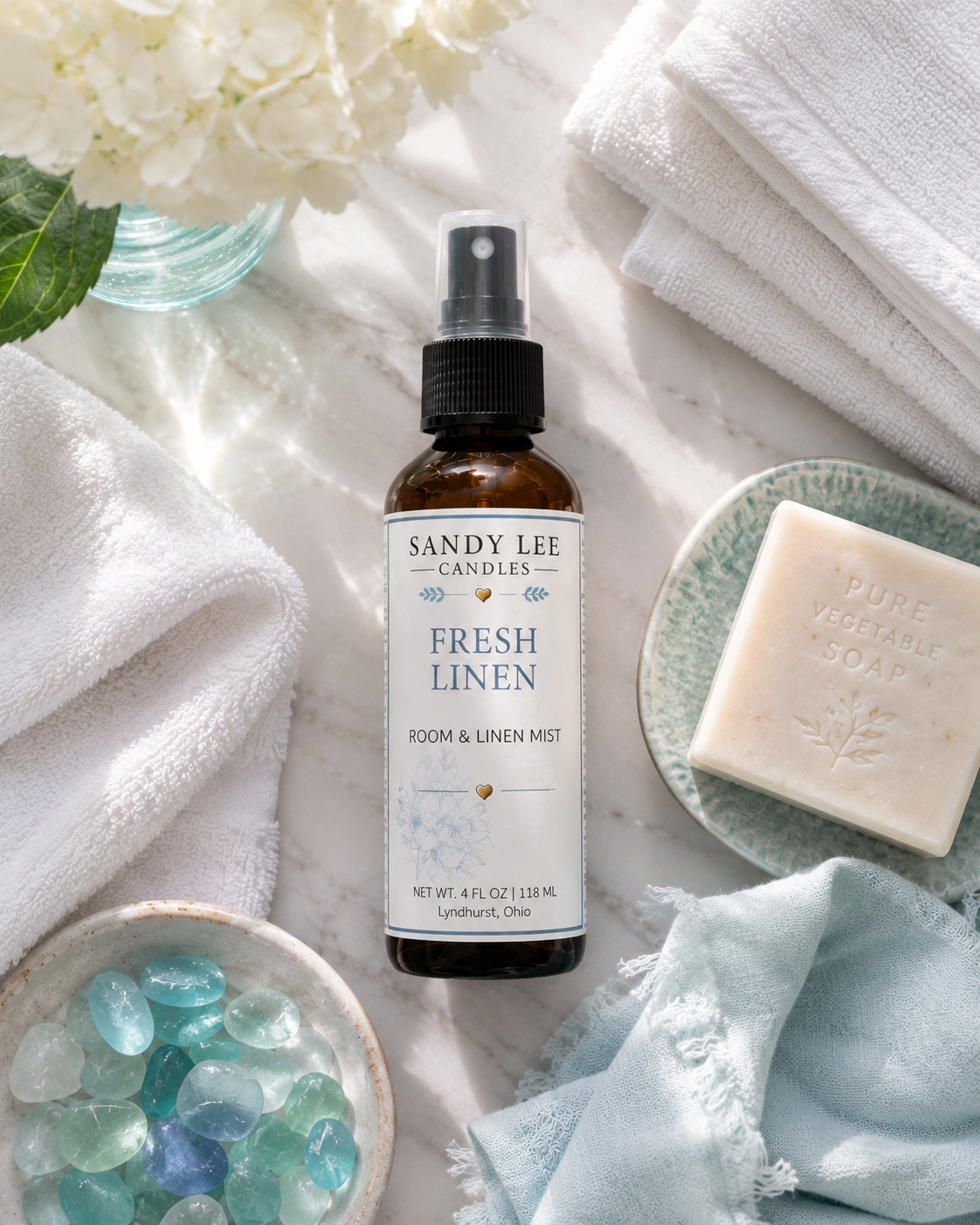 Fresh Linen Luxury Room & Linen Mist by Sandy Lee Candles in a premium amber glass bottle surrounded by crisp white linens, hydrangeas, and soft turquoise accents in a clean, luxury home setting.