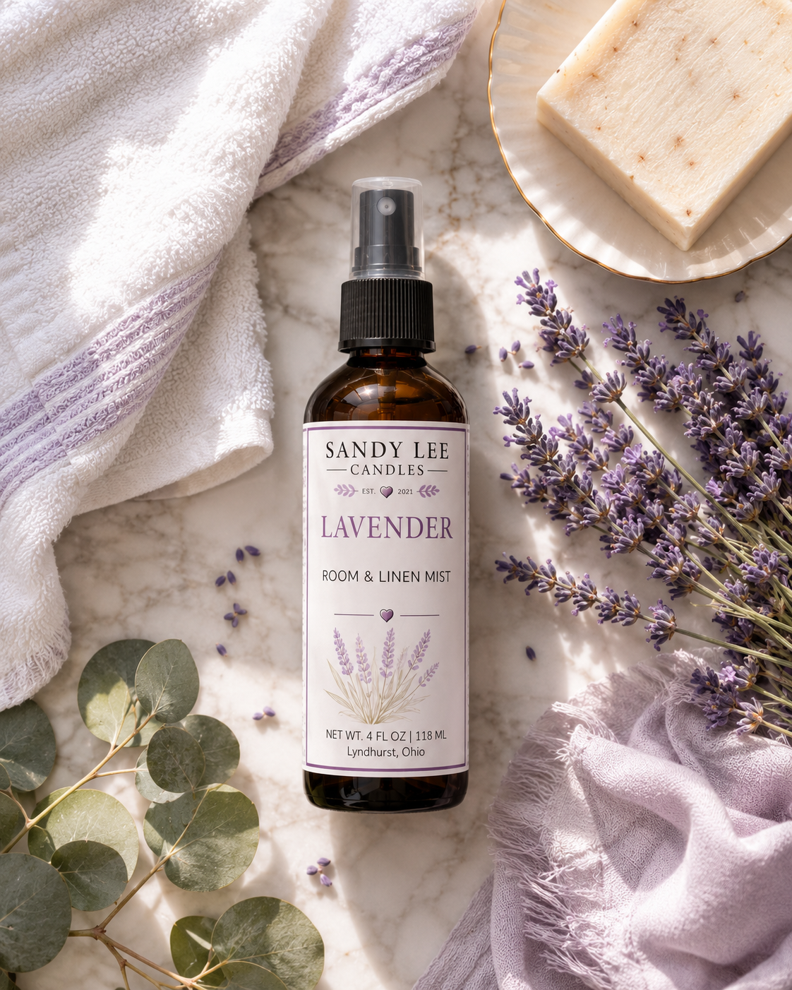 Sandy Lee Candles Lavender Luxury Room & Linen Mist in an amber glass bottle with fresh lavender, eucalyptus, and white hydrangeas in a spa-inspired setting.