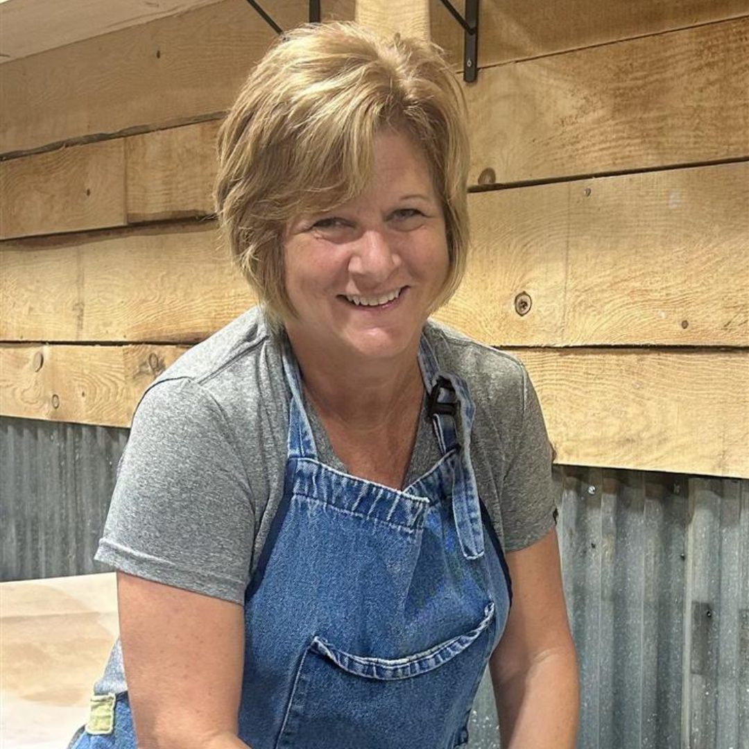 Sandy Lee wearing a blue apron sitting against a wooden wall making candles