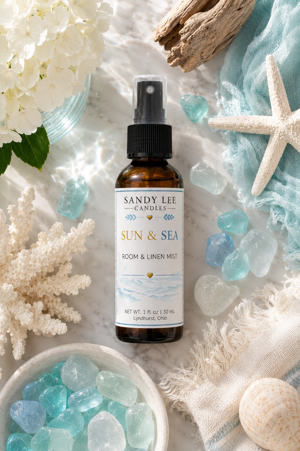 Sandy Lee Candles Sun & Sea Luxury Room & Linen Mist in an amber glass bottle styled with sea glass, white coral, driftwood, hydrangeas, and coastal décor in a bright ocean-inspired setting.
