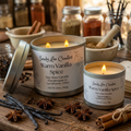 Two 'Sandy Lee Candles Warm Vanilla Spice' candles on a wooden surface with spices and jars in the background.