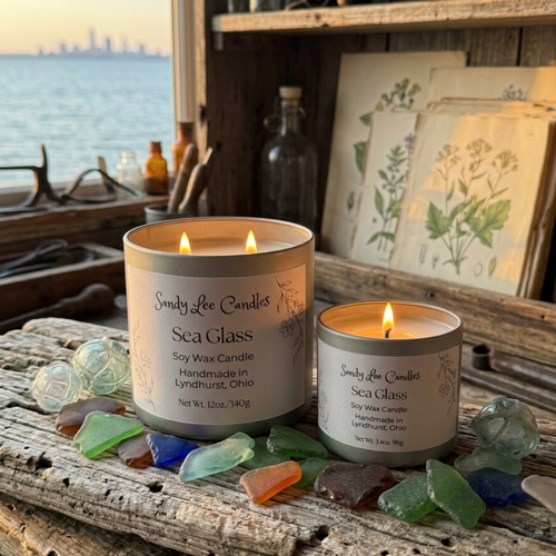Two lit candles from Sandy Lee Candles on a wooden surface with sea glass and a coastal background.